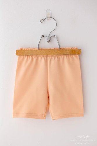 Dívčí šortky Lily Grey v jemné broskvové barvě zavěšené na dřevěném ramínku s kovovým hákem. 